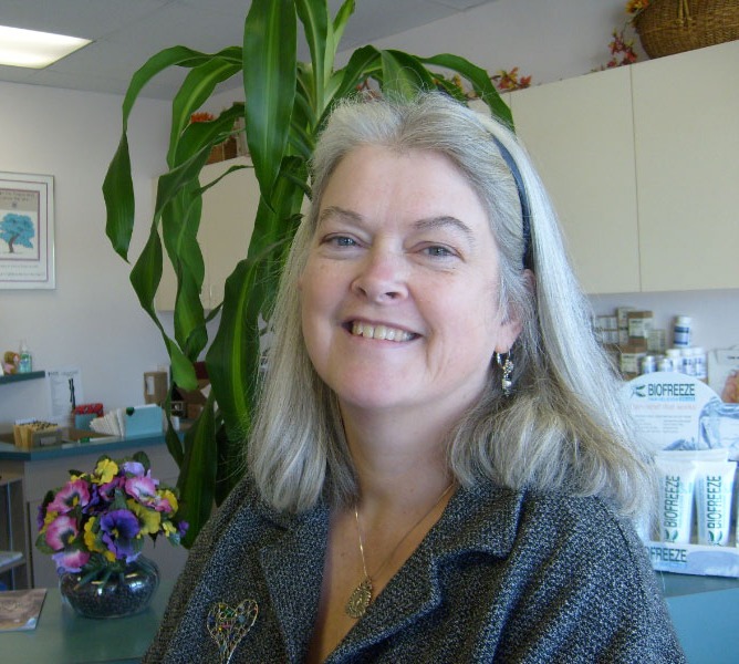 Dr. Mary Jane Doyle smiling in her office, with a vase of pansies and a tall corn plant behind her